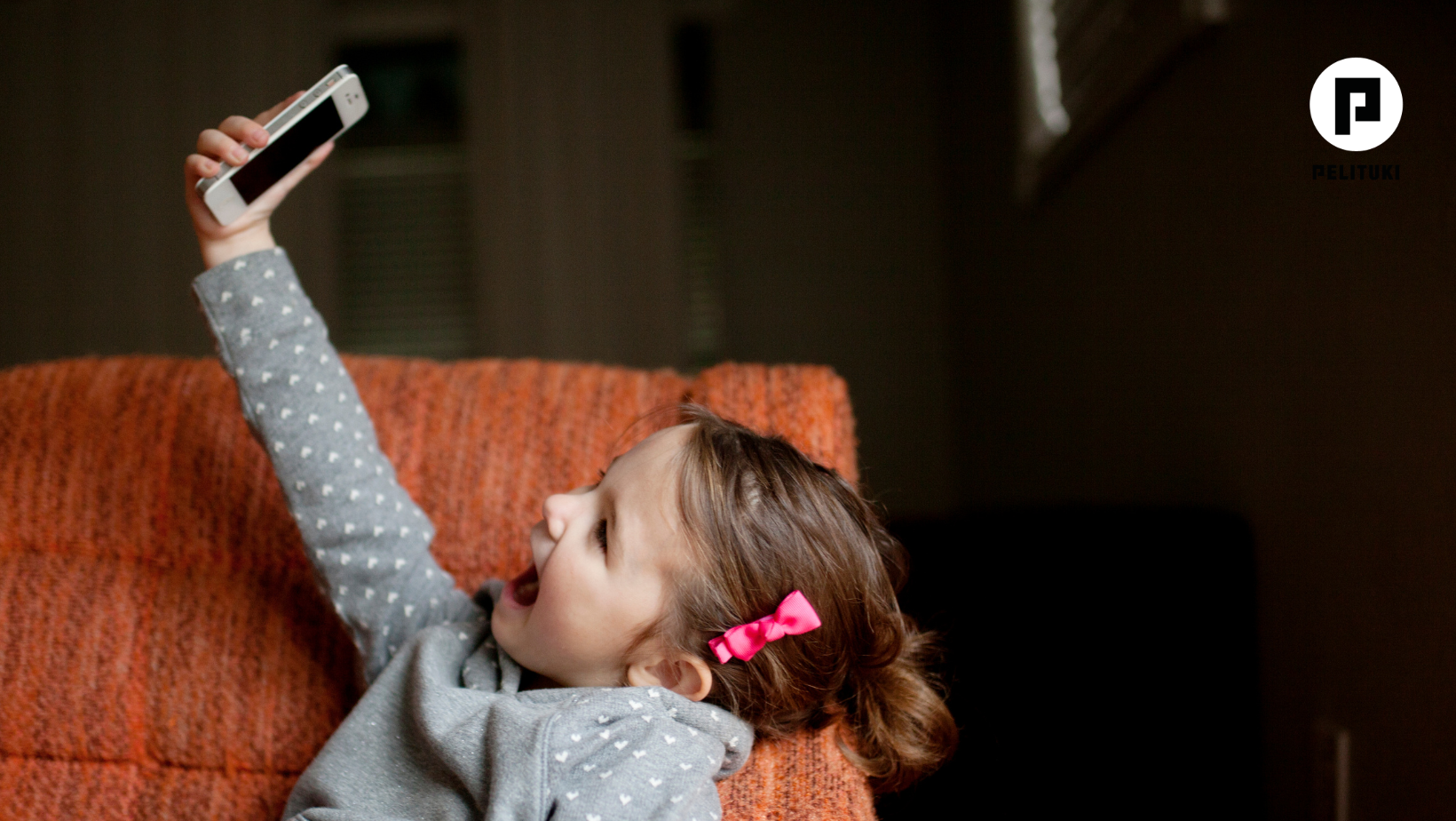 Pieni tyttö lepää sohvalla ja pitelee kädessään korkealla älypuhelinta, jota kohti hän katsoo iloisena